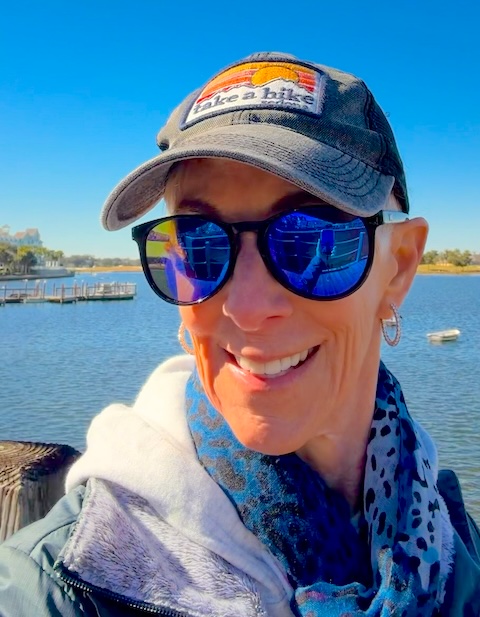 woman wearing sunglases, hat, scarf and jacket by the water